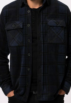 Mężczyzna ubrany w ciemnoniebieską i czarną koszulę w kratę typu button-up z klapkowymi kieszeniami na piersi, ręce w kieszeniach, pod spodem czarna koszulka henley.