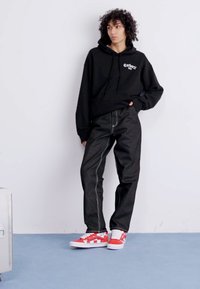 Černá mikina s logem, nahoře posazené tmavé džíny s kontrastním prošíváním a červené kostičkované slip-on boty na modelce před jednolitým pozadím.