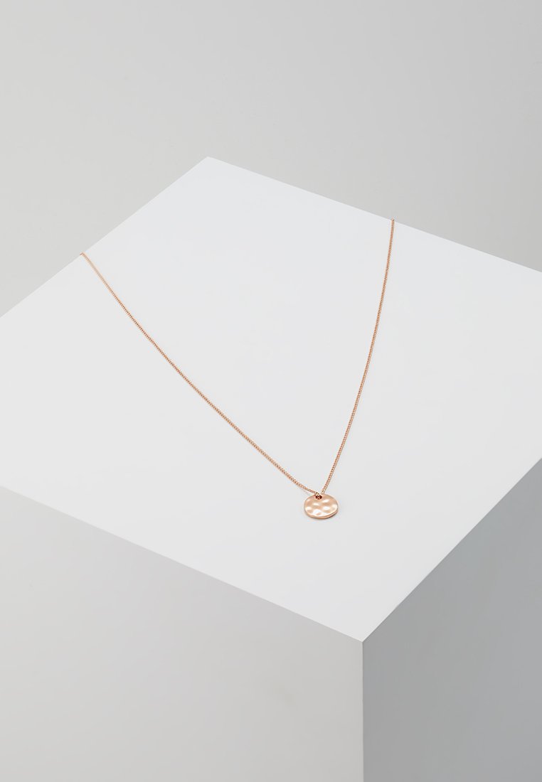 Collier en or rose avec un pendentif circulaire texturé, suspendu à une fine chaîne, présenté sur un support géométrique minimaliste.