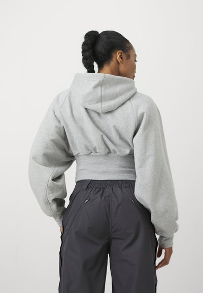 Mujer con coleta trenzada que lleva una sudadera corta gris y pantalones negros de talle alto, de pie de espaldas contra un fondo liso.