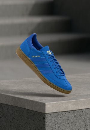 Blauwe Adidas Spezial sneaker met gumzool die boven een betonnen richel zweeft tegen een vervaagde achtergrond van grijze trappen.