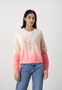 Maglione in gradiente rosa con logo a triangolo di Guess e un punto interrogativo, maniche lunghe e texture morbida, abbinato a jeans blu di denim.