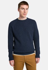 Maglione lavorato a maglia blu navy con una texture a costine, scollo rotondo e maniche lunghe. Indossato sopra una camicia chiara e abbinato a pantaloni beige.