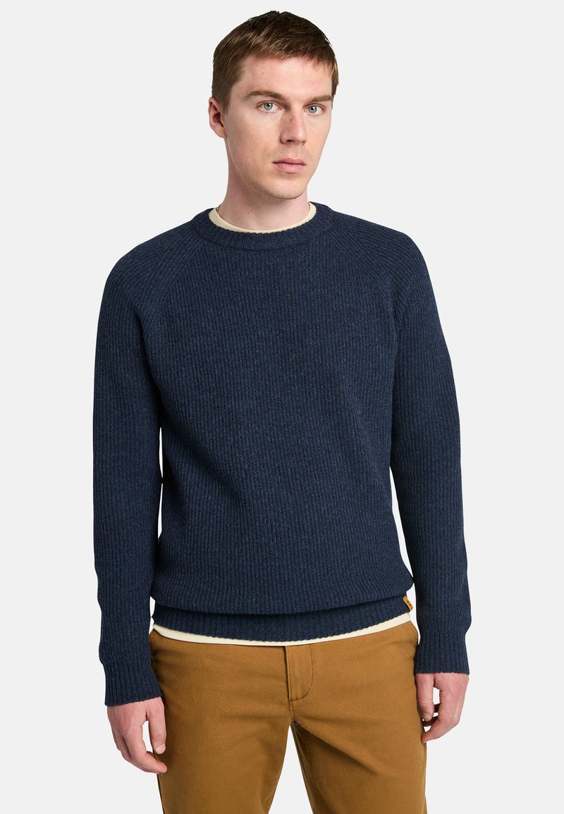 Maglione lavorato a maglia blu navy con una texture a costine, scollo rotondo e maniche lunghe. Indossato sopra una camicia chiara e abbinato a pantaloni beige.