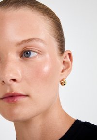 Χρυσό σκουλαρίκι με γυαλιστερή φινίρισμα που διαθέτει σχήμα δάκρυ. Το μοντέλο έχει λεία υφή δέρματος και διακριτικό μακιγιάζ.