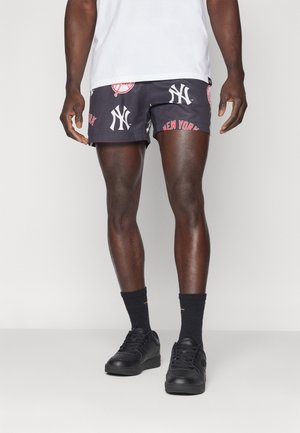 Mann trägt schwarze und weiße Shorts mit New York Yankees-Aufdruck, schwarze Socken, schwarze Turnschuhe und ein weißes T-Shirt vor einfachem Hintergrund.