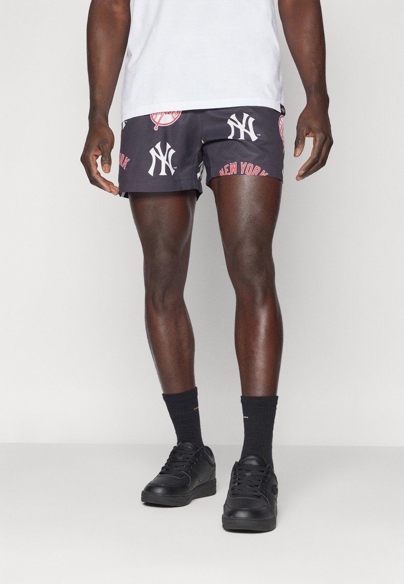 Homme portant un short imprimé New York Yankees noir et blanc, des chaussettes noires, des baskets noires et un t-shirt blanc sur un fond uni.