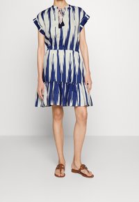 Flared dress in navy and cream with an ikat pattern, short sleeves, tie neck detail, and a ruffled hem. Worn with brown flat sandals.