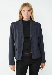 Navy pinstripe blazer with a tailored fit, featuring notched lapels, four buttons, and side pockets. Worn over a black top.