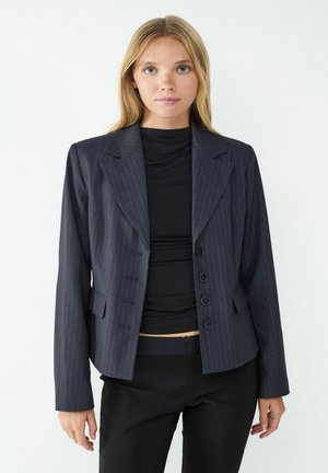Blazer en pinstripé marine à coupe ajustée, avec revers à cran, quatre boutons et poches latérales. Porté par-dessus un haut noir.