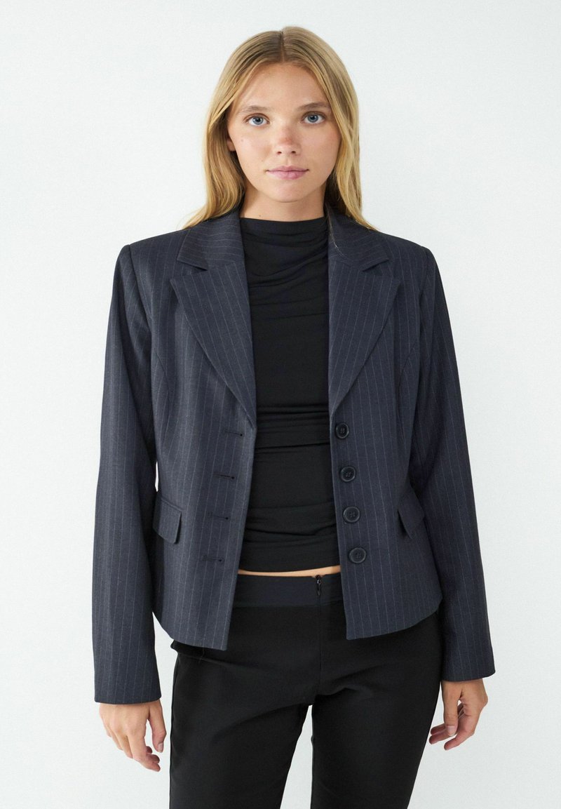 Navy pinstripe blazer with a tailored fit, featuring notched lapels, four buttons, and side pockets. Worn over a black top.