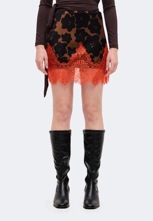 Modèle portant une mini-jupe à fleurs brunes et noires avec une bordure en dentelle orange vif, un haut sombre et des bottes noires jusqu’aux genoux.