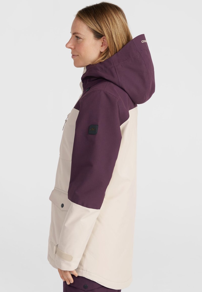 Lila und beige Jacke mit Kapuze, ausgestattet mit einem Frontreißverschluss, seitlichen Taschen und strukturierter Stoff. Akzente umfassen ein Logo auf dem Ärmel.