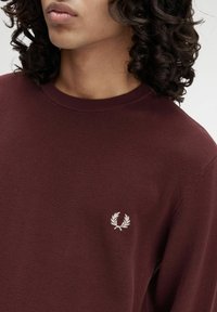 Persona con capelli ricci che indossa un maglione corto testurizzato color bordeaux, caratterizzato da un piccolo emblema di alloro bianco sul petto.