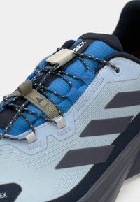 Αθλητικό παπούτσι σε ανοιχτό μπλε χρώμα με σκούρες λεπτομέρειες, υφή στην επιφάνεια, σύστημα γρήγορου δεσίματος και έντονες μαύρες ρίγες. Κατάλληλο για εξωτερική χρήση.
