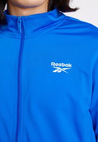 Casaco azul com fecho éclair, feito de um tecido suave e leve. Apresenta um colarinho alto e o logótipo da Reebok em branco na área do peito esquerdo.