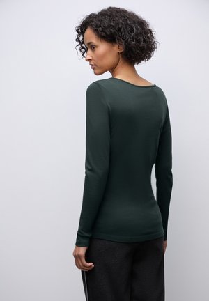 Maglietta a maniche lunghe di un verde scuro con scollatura a barca e design aderente, abbinata a pantaloni grigi. Texture liscia, dettagli minimali.