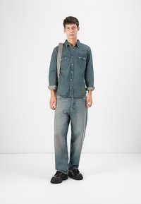 Dunkelblaue Denim-Hemd mit zwei Brusttaschen und Knopfleiste. Getragen mit hellblauen weiten Jeans und klobigen schwarzen Schuhen.