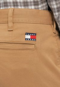 Κοντινή λήψη στην τσέπη μπεζ παντελονιού με το λογότυπο Tommy Jeans; μέρος ενός καρό πουκαμίσου φαίνεται στη μέση.