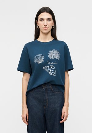 Γυναίκα με μακριά σκούρα μαλλιά, που φορά χρυσούς κρίκους αυτιών, μπλε κοντομάνικο μπλουζάκι με λευκά κοχύλια και τη λέξη "Weekend", συνδυασμένο με σκούρα τζιν παντελόνια.