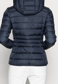 Giacca imbottita blu navy con cappuccio, caratterizzata da un design a righe orizzontali e una texture liscia. Indossata con jeans bianchi, che mettono in evidenza una silhouette aderente.