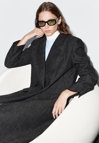 Šedý přizpůsobený oblek s texturovanou tkaninou, s dvojitým stylem a širokými klopami. Černé sluneční brýle a bílý rolák zvýrazněný.