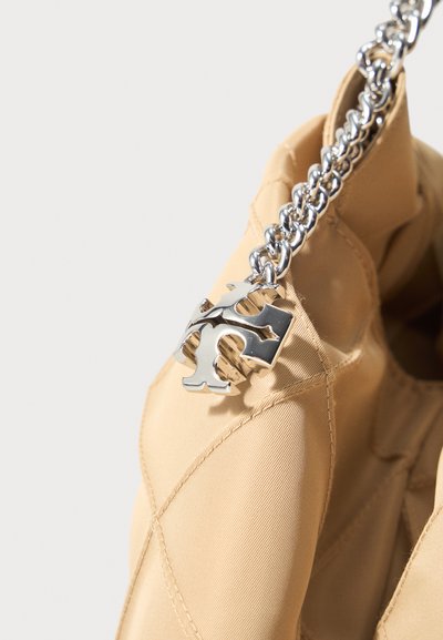 Κοντινή όψη μπεζ τσάντας από καπιτονέ ύφασμα με ασημένια αλυσίδα λουρί και διακοσμητικό ασημένιο λογότυπο ως γούρι.