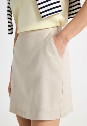 Kvinde iført beige A-formet nederdel med hånden i lommen, kombineret med gul top og stribet sweater hængende over skuldrene.