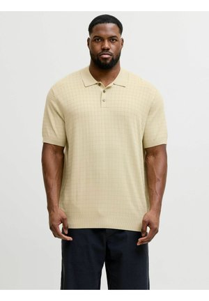 Homme debout face à l’avant portant un pull en maille beige à manches courtes avec col, avec un motif à carreaux subtil, et un pantalon foncé.