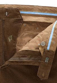 Intérieur en tissu brun de pantalons avec une poche zippée, un bouton et une broderie de feuille verte. Doublé d'un motif géométrique subtil.