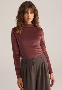 Moteris su garbanais plaukais, vilkinti ilgu rankoviu bordo spalvos marškinį ir aukšto liemens rudas klostuotas kelnes, prieš paprastą foną.