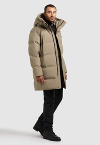 Abrigo acolchado beige con capucha, con un diseño acolchado, bolsillos con cremallera y corte recto, acompañado de jeans negros y botas.