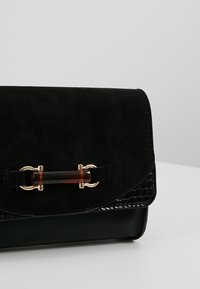 Pochette en cuir noir et daim avec une fermeture à barre en écaille et ton or, posée sur une surface blanche unie.