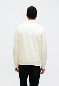 Krém színű pulóver texturált anyagból, bordázott mandzsettákkal és szegéllyel. Gallérral és kényelmes szabással rendelkezik, hátulnézetben látható.