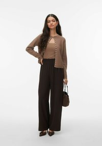 Cardigan marron à boutons en perles, associé à un débardeur marron et un pantalon marron à jambes larges. Le mannequin tient un petit sac marron.