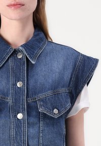 Denim ilmanhiha shirt, jossa on terävä kaulus, hopeiset napeilla ja kaksi rintataskua. Kangas on sininen, jossa on hieno tekstuuri.