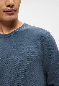 Blå strikkegenser med rund hals og teksturert mønster. Har et subtilt preget logo på brystområdet.