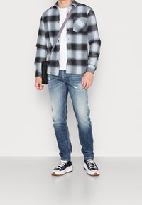 Camicia a quadri nera e blu in flanella, t-shirt bianca sotto, jeans azzurri strappati e sneakers nere con suola bianca e dettagli beige.
