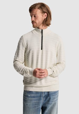 Homme aux cheveux châtain clair portant un pull crème texturé à demi-fermeture éclair et un jean bleu, debout, les mains jointes, regardant vers la gauche.