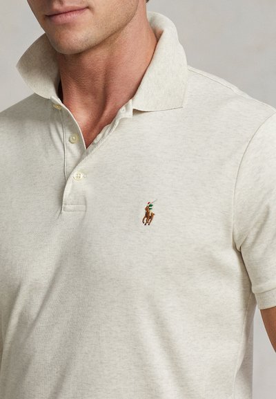 Mies, joka käyttää vaalean beigeä poolopaitaa, jossa vasemmassa rinnassa on kirjailtu logo hevosella ratsastavasta poolopelaajasta.