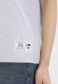 Camiseta de algodón gris con un parche de logo rectangular con acentos en azul, rojo y blanco, que indica "Tommy Jeans, Nueva York."