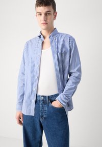 Blue and white striped button-up shirt with a collar and pocket, worn over a white tank top, paired with blue jeans.
