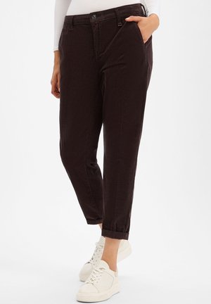 Woman wearing dark brown cropped pants with cuffed hems and white sneakers, standing with one hand in pocket.