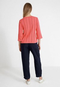 Korall színű kötött felső, 3/4-es ujjal, függőleges csíkokkal és nyitott szemcsemintázattal; navy színű bőszárú nadrággal és fehér sportcipővel párosítva.