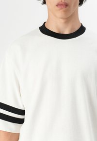 Hvid kortærmet t-shirt med en rund sort krave og to sorte striber på hver ærme. Stoffet ser blødt og glat ud.