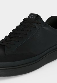 Μαύρο δερμάτινο sneaker χαμηλού τύπου με μαύρα κορδόνια, υφασμένο πάνω μέρος και λογότυπο "UGG" στη γλώσσα, εμφανιζόμενο από γωνία.