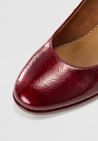 Escarpin en cuir texturé rouge avec un bout arrondi, doté d'une finition brillante et d'un talon légèrement surélevé. L'intérieur est beige avec des coutures visibles.