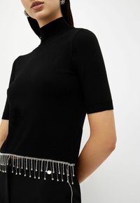 Vrouw in een zwart turtleneck top met korte mouwen, versierd met zilveren kristallen franjes aan de onderkant, en zwarte broek met kristalaccenten.