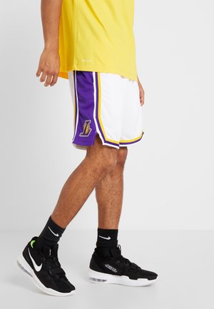 Mężczyzna w żółtej koszulce, białych i fioletowych spodenkach do koszykówki z logo Lakers, czarnych butach Nike i czarnych skarpetkach Nike, stoi na szarej podłodze.