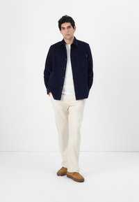 Chaqueta de cordón marina con cierre de botón, camisa gris clara, pantalones crema y zapatos color tostado. Presenta bolsillo en el pecho y corte relajado.